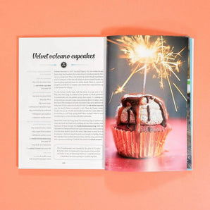 The Cake Book by Crumbs & Doilies open to a recipe page with a sparkler-topped velvet volcano cupcake photo.